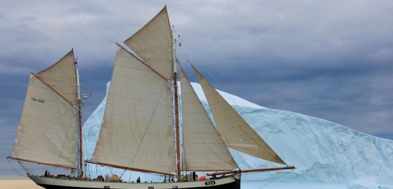 Traditional sailing ship Tecla will all her sails up, passing a large white iceberg in front of a moody sky. Polar exploration sailing adventure holidays with Classic Sailing.