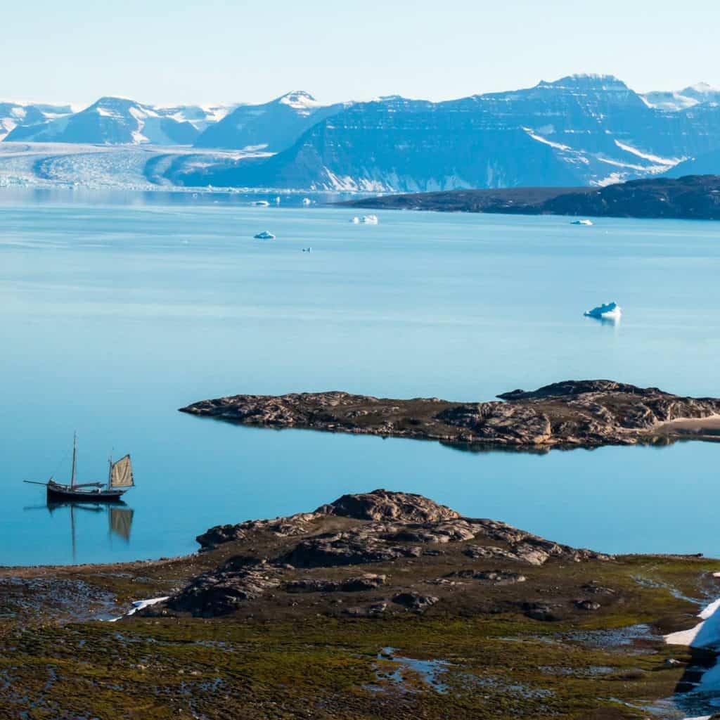 Tall ship Tecla anchored in Greenland. An expanse of water with distant icy mountains and a green foliage in the foreground. Enjoy polar sailing holidays with Classic Sailing.