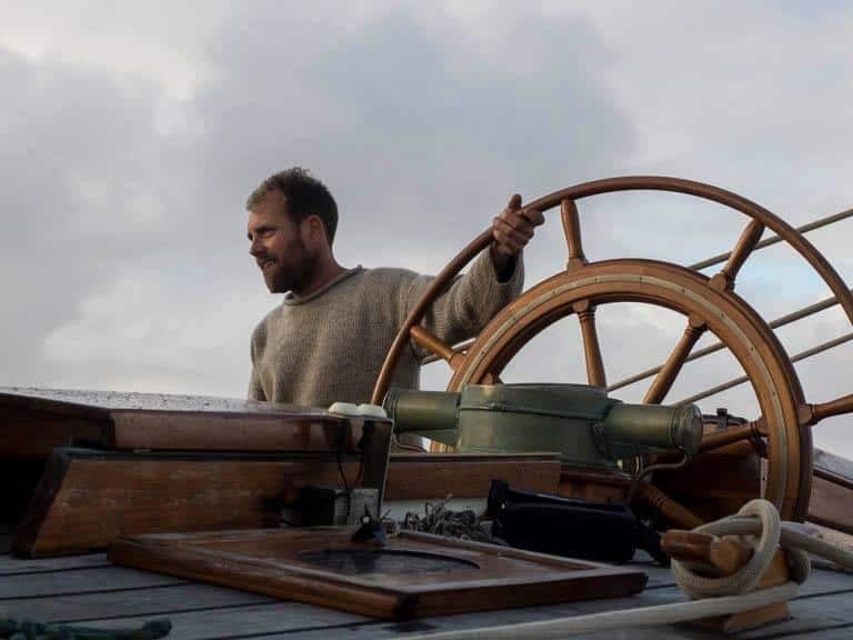 Crew member at the wheel of Tecla wearing a woolly jumper. Experience sailing in high latitudes with adventure holidays from Classic Sailing