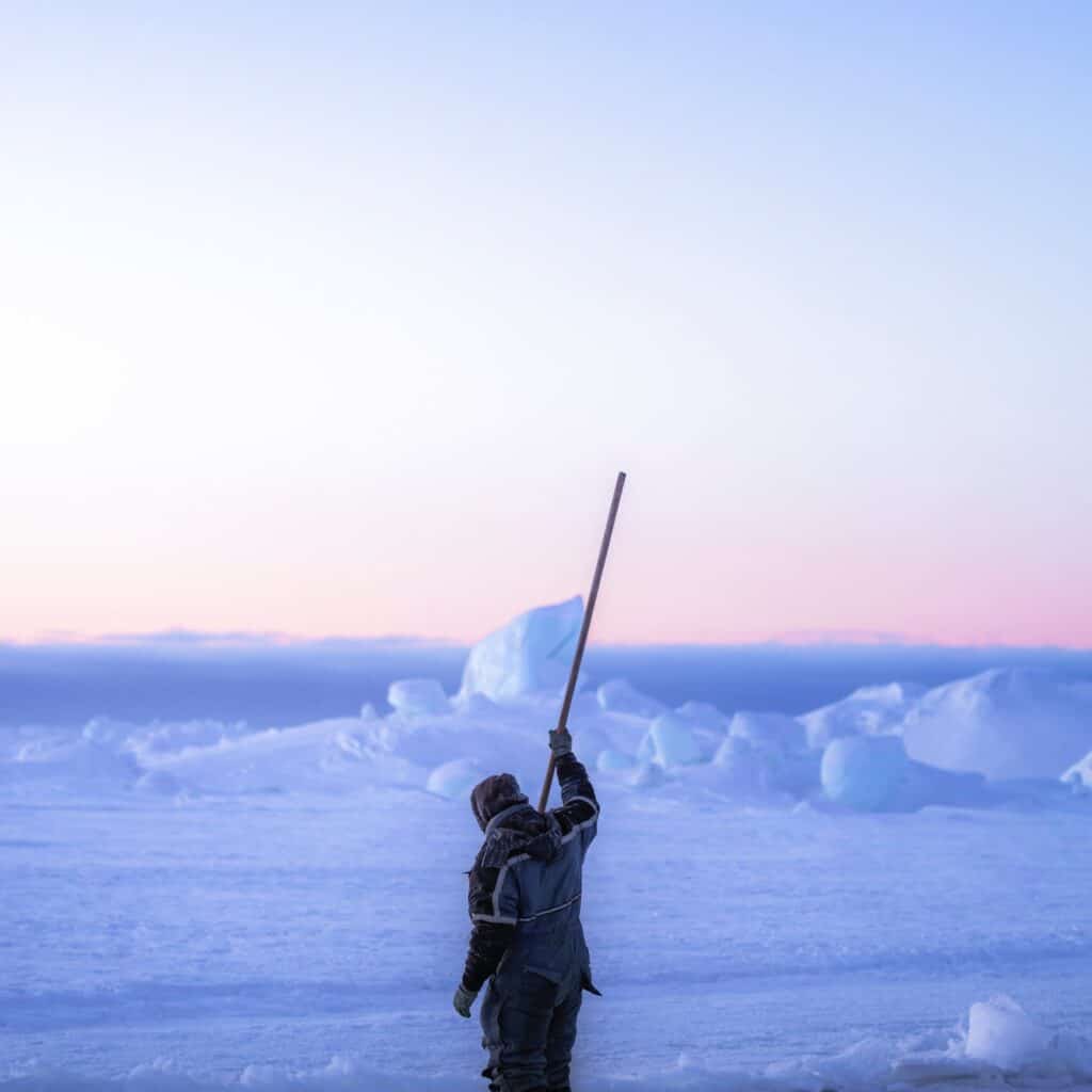 Inuit spear fishing on the ice, with a bright white sky in the background. Polar sailing holidays with Classic Sailing.