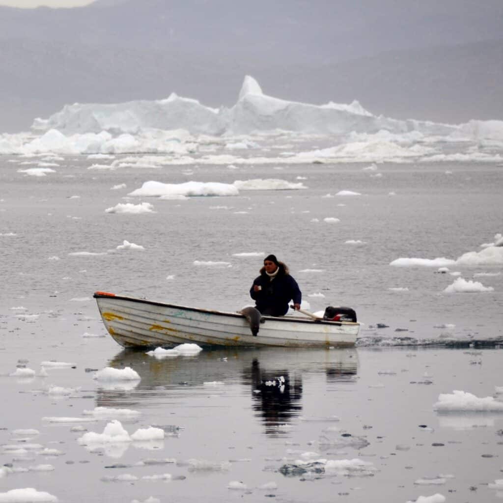 Inuit seal hunter in a small boat, steering through icy grey water. Arctic Greenland sailing adventures with Classic Sailing.
