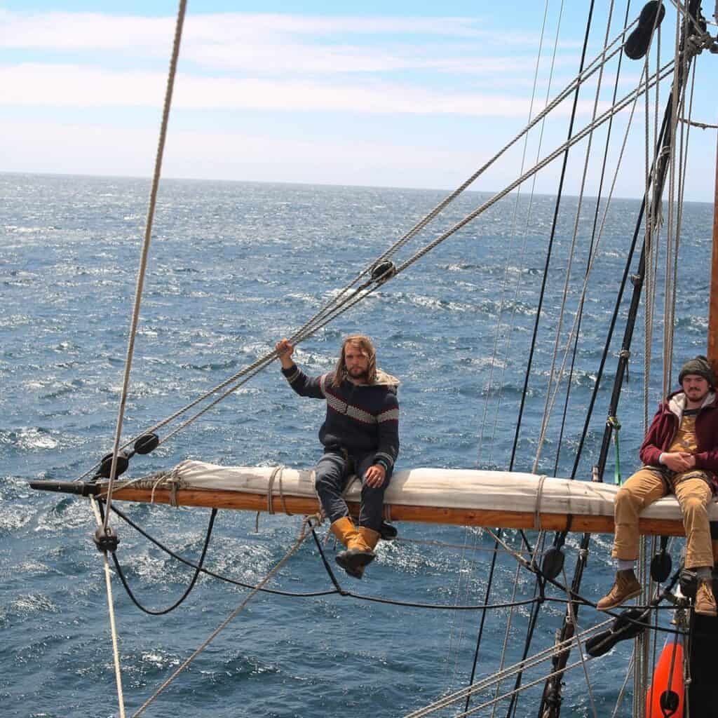 Brig Phoenix skipper Joel sat on the yardarm up the rigging with a grey blue sea below. Tallship crew up the rigging. Square sails. Adventure sailing holidays with classic sailing