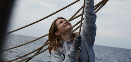A female sailor hauling lines on the deck of Lugger Grayhound, with a smile on her face. You can feel the same with a sailing adventure through Classic Sailing.