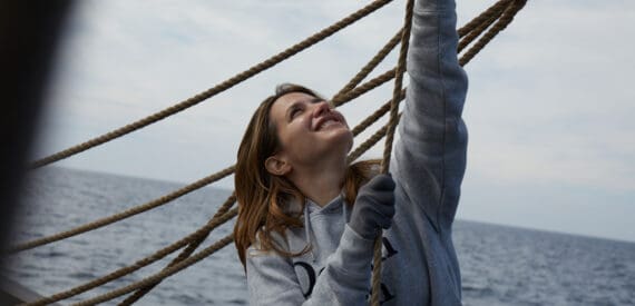 A female sailor hauling lines on the deck of Lugger Grayhound, with a smile on her face. You can feel the same with a sailing adventure through Classic Sailing.