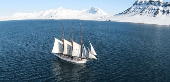Linden on a sailing and scientific expedition in Svalbard. Sailing in the sunshine with a snowy landscape behind. Join her through Classic Sailing