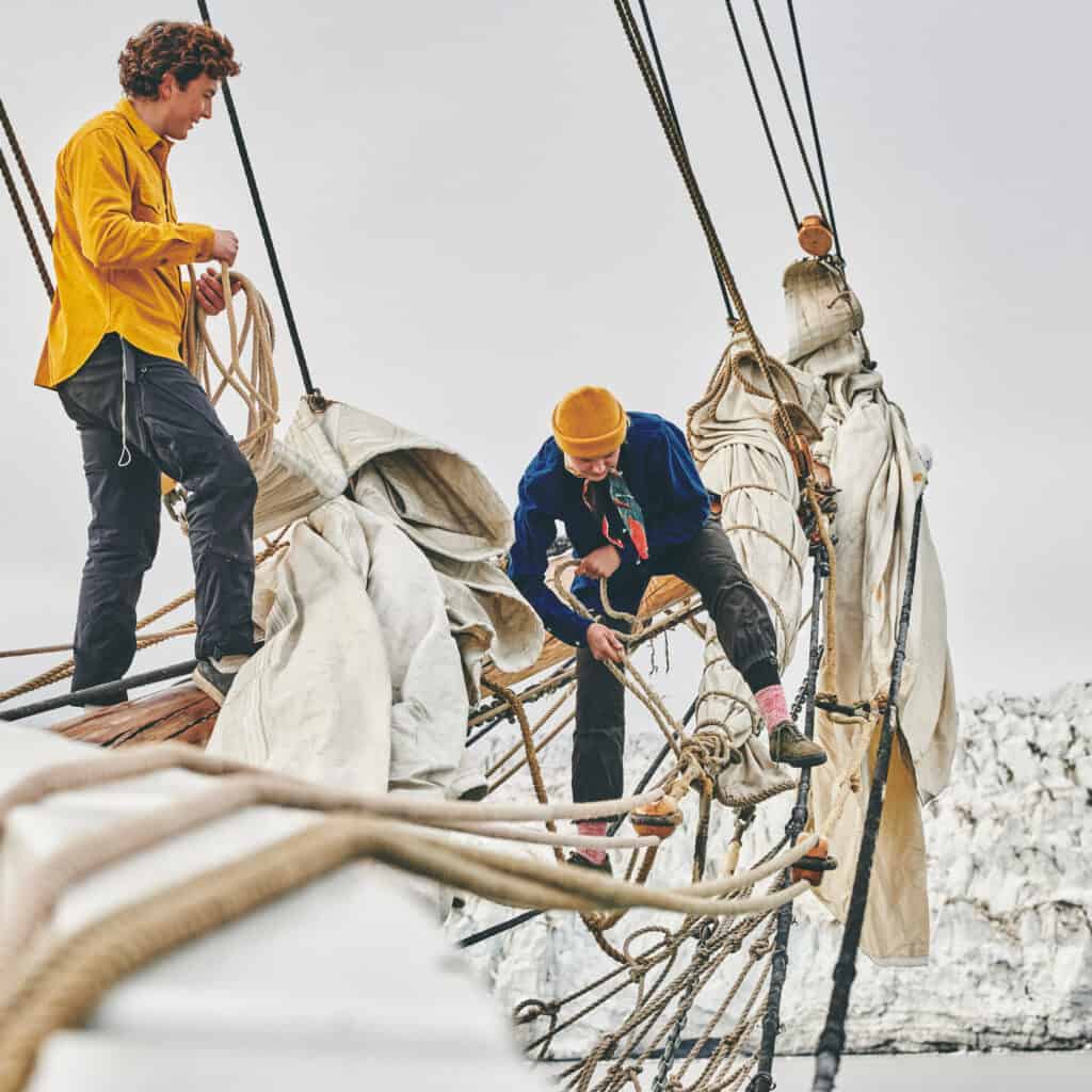 Crew on Linden furl sails on the bowsprit, with a glacier in the background. Join the ship for an arctic expedition to svalbard through Classic Sailing.
